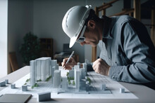 Generative AI Illustration Of Side View Male Architect In Hard Hat And Sunglasses Sitting At Table Near Mockup Of New Modern Residential Complex And Correcting Drawing While