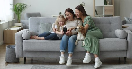 Wall Mural - Three generation of women and dog relax on sofa in comfortable living room, use modern cellphone to watch funny videos, on-line content, smile enjoy new filter, spend nice time with family on weekend