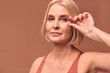 © Velista production - A beautiful blonde older woman on a beige background shows wrinkles near her eyes. Skin aging and skin care concept.