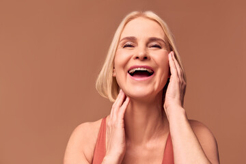 Wall Mural - Live photo of a happy older woman laughing and touching her face with her hands while posing in front of the camera. Woman's beauty, facial skin care.Beige background.