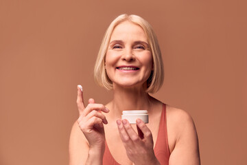 Wall Mural - Close-up portrait of a happy beautiful blonde older woman holding a jar of cream in one hand and cream on her finger in the other. Woman laughing and looking at camera on beige background.