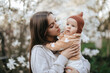 © VikaNorm - A teenage girl with long hair in a gray sweatshirt is holding her newborn sister in a garden with blooming white magnolia.