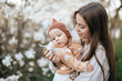 © VikaNorm - A teenage girl is showing her newborn sister a flower from a magnolia tree in a blooming garden.