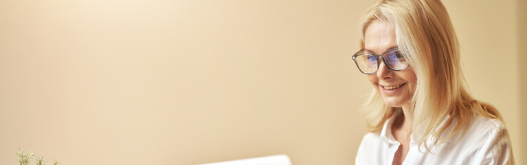 happy mature businesswoman in glasses smiling while using laptop, working in the office