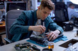 © Roman - confident guy repairing parts in disassembled broken laptop using screwdriver and different tools on table in workshop, professional technician at work place, concentrated on repair