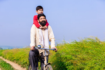 Wall Mural - Rural Indian father and son riding bicycle in agricultural field in bright sunny day, Parents with son, Copy space.