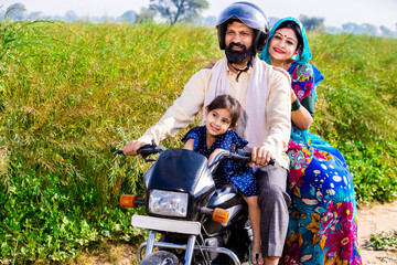 Wall Mural - Happy young rural Indian family wearing safety helmet riding on motorcycle in village agricultural field. Mode of transportation, Copy space