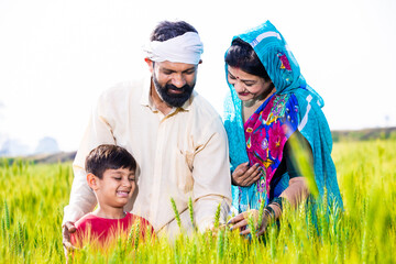 Wall Mural - Portrait of happy young indian farmer family touching wheat crop standing at agriculture field in bright sunny day. Parent with child ,Rural india concept.