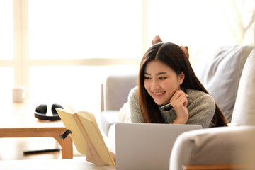 Young business freelance Asian woman working on laptop checking social media while lying on the sofa when relax in living room at home.