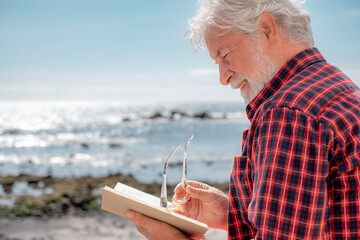 Wall Mural - Portrait of senior bearded man reading a book in front to the sea. Elderly relaxed pensioner with eyeglasses enjoying retirement in outdoor