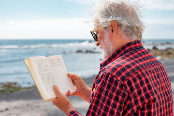 Wall Mural - Back view of senior bearded man reading a book in front to the sea. Elderly relaxed pensioner with sunglasses enjoying retirement in outdoor