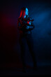 © Вероника Преображенс - A young beautiful woman with long hair in a black leather jacket is holding a gun in the dark at night. Robber in the city in neon blue and red light.