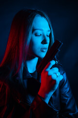 A young beautiful woman with long hair in a black leather jacket is holding a gun in the dark at night. Robber in the city in neon blue and red light.