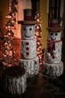 Snowmen constructed from tree logs, wearing hats, standing next to illuminated Christmas trees
