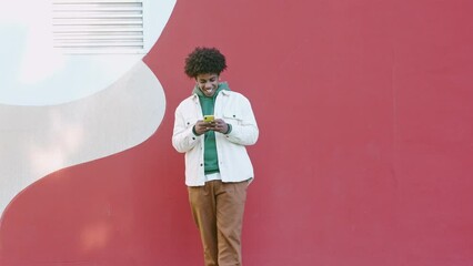 Wall Mural - Cool young African American guy holding mobile phone standing at bright urban city wall. Happy stylish hipster teenager using cell outdoors, looking at cellphone texting message in social media chat.