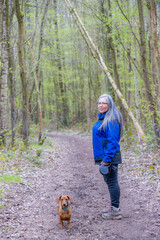 Wall Mural - Senior adult hiker standing in profile on trail next to her short-haired brown dachshund, wooded landscape in background, long gray hair, Strijthagerbeekdal Nature Reserve, South Limburg, Netherlands