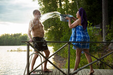 Aged Couple Having Fun After Finnish Sauna On Wooden Cottage Pier In A Lake. Mature Woman Pouring Cold Water From Basin Over Her Partner. Typical Finnish Summer. 