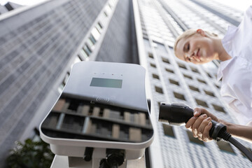 Fisheye view focus hand holding, inserting, plugging ev charger from public charging station for electric vehicle with blurred progressive businesswoman and residential city building in background.