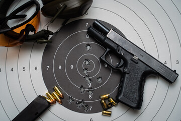 Shooting from a weapon at a target in a shooting range.  A pistol, goggles and earmuffs, and a target with bullet holes in the center.