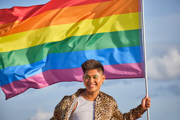 gender non binary asian person from front holding a rainbow gay pride flag blowing in the wind