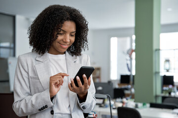 Young smiling successful African American business woman, beautiful female entrepreneur businesswoman using smartphone, cellphone application, online communication, standing in modern office indoors.