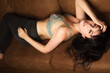 © Angela - Portrait of a caucasian model in her 30s wearing a blue bra and dark high waisted jeans while lying on a brown couch. She has dark brown hair and blue eyes.