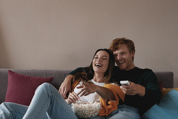 Wall Mural - Beautiful young couple watching TV and laughing while relaxing on the couch at home together