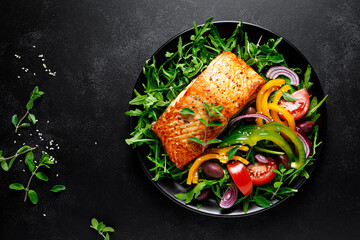 Canvas Print - Salmon fillet grilled and fresh vegetable green salad of arugula with tomatoes, olives and bell pepper on black background, healthy food, mediterranean diet, top view