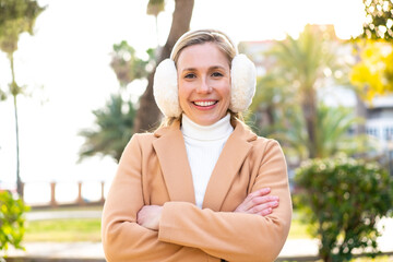 Wall Mural - Young blonde woman wearing winter muffs at outdoors keeping the arms crossed in frontal position