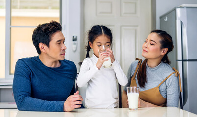 Portrait of enjoy happy love asian family father and mother with little asian girl smiling and having protein breakfast drinking and hold glasses of milk at table in kitchen