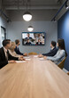 Team of businesspeople meeting at table in small conference room, discussing work project with remote diverse colleagues on group video call, turning heads to screen with facial head shots