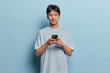 © wayhome.studio  - Horizontal shot of handsome brunet Asian man holds smartphone in hands types text messages or surfs social networks dressed in casual t shirt isolated on blue background. People and technology concept