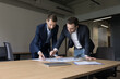 © fizkes - Two business partners in formal clothes standing at meeting table over paper marketing reports, reviewing statistic documents, discussing startup project plan, strategy, analyzing sales, risk