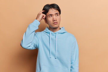 Young thoughtful young Asian man with dark hair scartches head tries to remember something has doubtful expression gathers with thoughts dressed in casual blue sweatshirt isolated on brown background