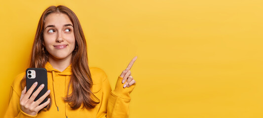 Horizontal shot of good looking teenage girl points fore finger at upper left corner has news about bargains holds modern smartphone in hand wears sweatshirt isolated over vivid yellow background