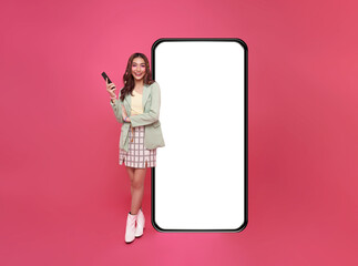 happy asian woman using mobile phone standing at big giant smart phone with empty white screen, pres