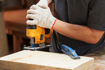 the hands of a Caucasian worker in white gloves are sawing with an electric jigsaw with laser illumination a wooden blank fixed with a clamp