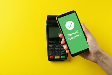 Woman using terminal for contactless payment with smartphone on yellow background, top view. Transaction completed screen on device