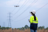 Fototapeta Storczyk - A female electrical engineer fielding wearing vr goggles using drone to inspect power station to view power plant planning work, high voltage transmission towers in aerial view.