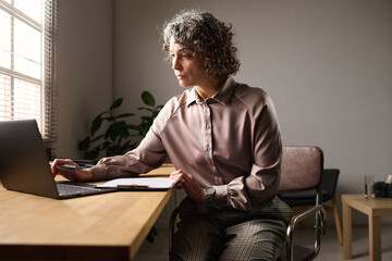 Leinwandbilder - Elegant mature female psychologist sitting by workplace in front of laptop while looking through personal data of patients and making notes