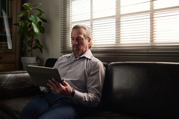 Poster - Confident mature psychologist looking at tablet screen during online session with patient while sitting on black leather couch by window
