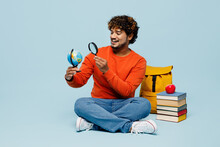 Full Body Young Teen Indian Boy Student Wear Casual Clothes Sit Near Books Backpack Bag Hold Earth World Globe Use Magnifier Isolated On Plain Blue Background. High School University College Concept.
