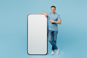 Wall Mural - Full body young happy man wear casual t-shirt point index finger on big huge blank screen mobile cell phone with workspace copy space mockup area isolated on plain pastel light blue cyan background.