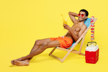 Full body young sexy man wear orange shorts swimsuit relax near hotel pool sit in deckchair drink citrus juice look camera isolated on plain yellow background Summer vacation sea rest sun tan concept