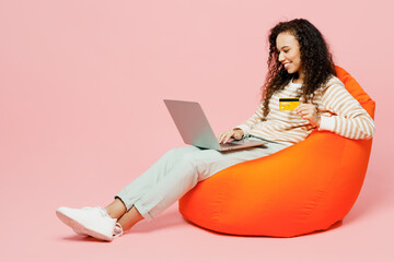 Wall Mural - Full body happy smiling young woman she wears casual clothes sit in bag chair using laptop pc computer credit bank card shopping online order delivery booking tour isolated on plain pink background.