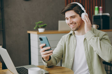 Wall Mural - Young happy man wear casual clothes headphones sits alone at table in coffee shop cafe restaurant indoors work or study on laptop pc computer use cell phone. Freelance mobile office business concept.