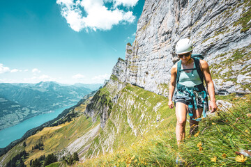 Wall Mural - Close up of woman hiking on flowery trail next to steep rock wall with scenic view on Walensee and Churfürsten mountain range in the background. Schnürliweg, Walensee, St. Gallen, Switzerland, Europe.