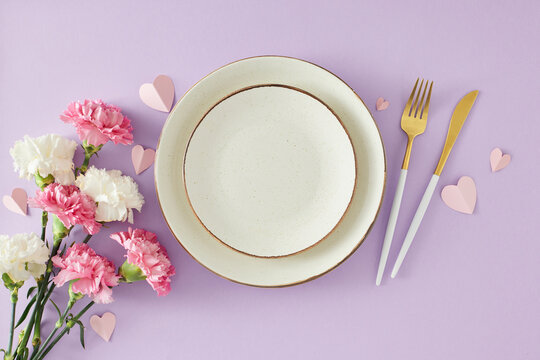 Mother's day table setting concept. Top view photo of empty plate and cutlery knife fork bunch of carnation flowers and paper hearts on lilac background. Flat lay with space for your greeting or text