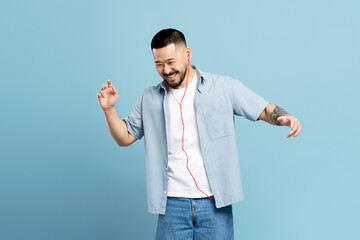 Attractive happy asian man with stylish tattoo, wearing headphones listening music, dancing isolated on blue background, copy space. Technology, positive emotions concept