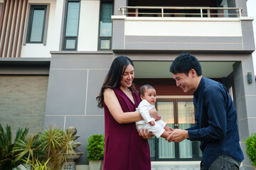 Wall Mural - happy family, cheerful father and mother holding and playing with newborn baby while standing in front of their house